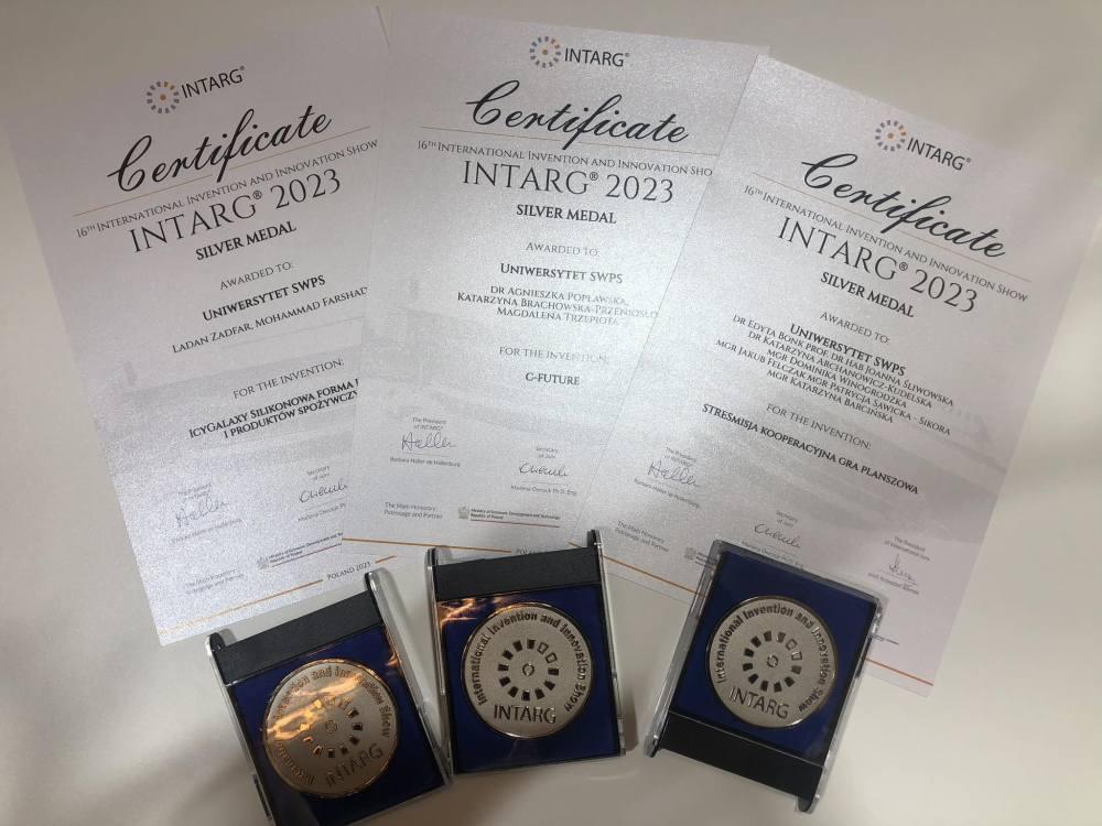 Zdjęcie trzech srebrnych medali i odpowiadających im certyfikatów dla projektów Uniwersytetu SWPS