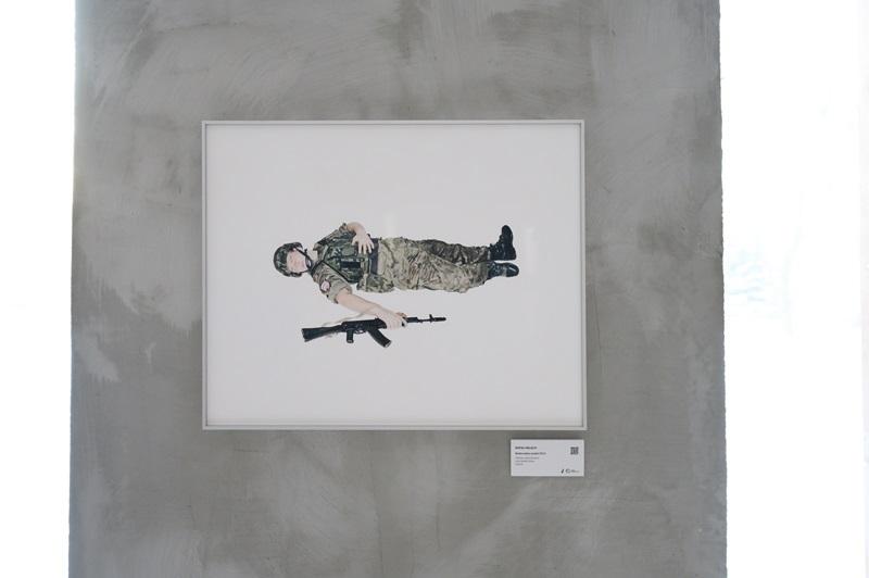 Fotografia autorstwa Rafała Milacha, przedstawiająca żołnierza leżącego na ziemi, z lewą ręką na klatce piersiowej i prawą na leżącej obok broni. 
