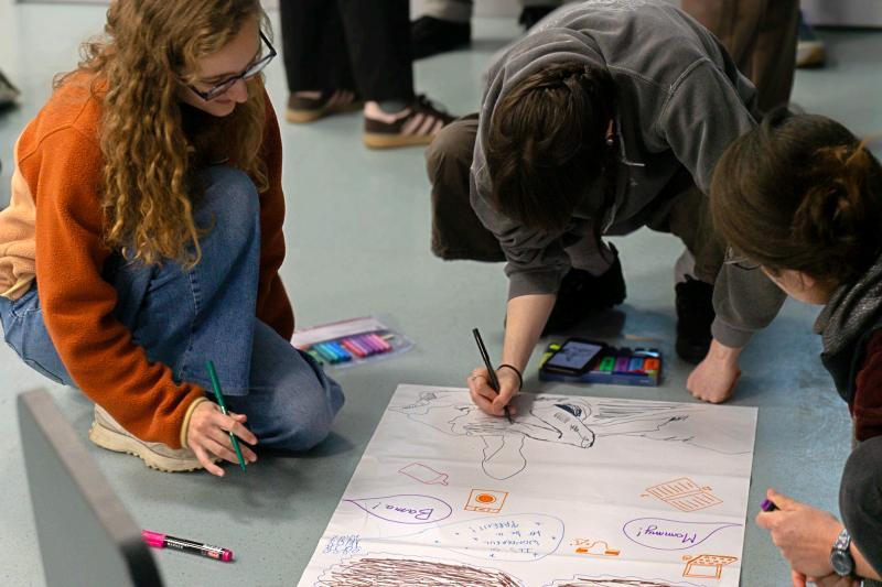 Troje studentów siedzi na podłodze i rysuje na dużej kartce papieru