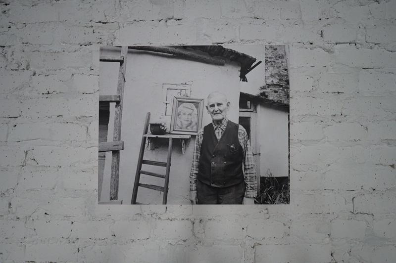 Fotografia autorstwa Zofii Rydet wyeksponowana na ścianie w czarnej ramie. Na zdjęciu widać starszego mężczyznę stojącego przed swoim domem. W tle widać kawałek drabiny i portret kobiety w ramie.