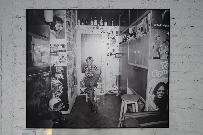 Fotografia autorstwa Zofii Rydet wyeksponowana na ścianie w czarnej ramie. Na zdjęciu widać młodego mężczyznę siedzącego na stołku w swoim pokoju. Na ścianach widać wiele plakatów, w tle stoi kolekcja butelek. Widać kask położony na stołku, oraz meblościankę i fotel.
