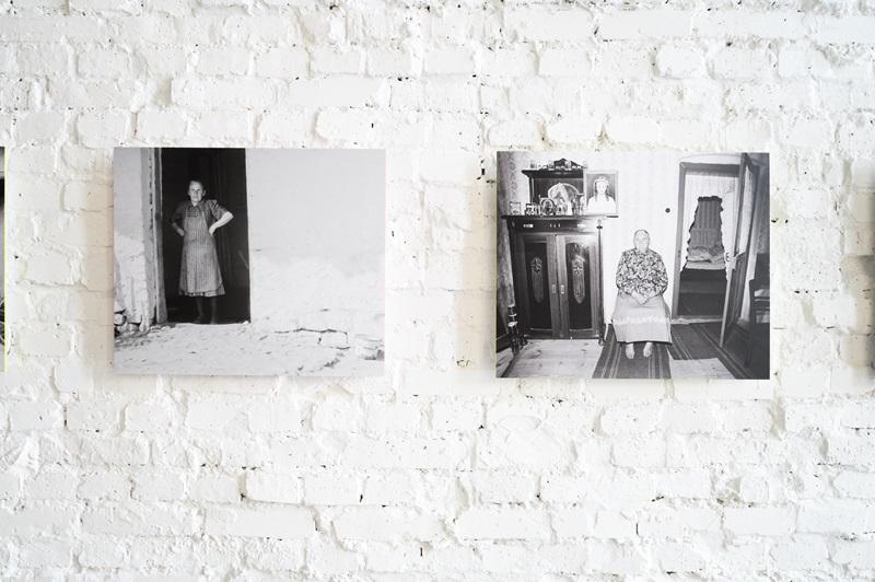 Dwie fotografie autorstwa Zofii Rydet wyeksponowane na ścianie. Na pierwszej widać starszą panią stojącą na ganku, na drugiej kobietę siedzącą na środku pokoju na krześle. W tle widać charakterystyczne koronkowe firanki, a krzesło na którym siedzi stoi na wzorzystej wykładzinie, obok masywnej drewnianej komody. 