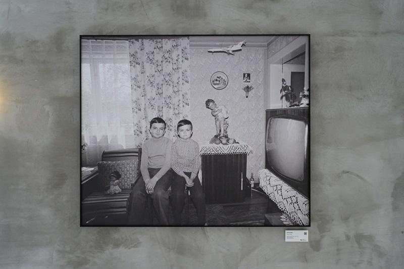 Fotografia Zofii Rydet wyeksponowana na ścianie w czarnej ramie. Na zdjęciu widać dwóch chłopców siedzących koło siebie na środku salonu, w którym widać mocno styl lat 70.