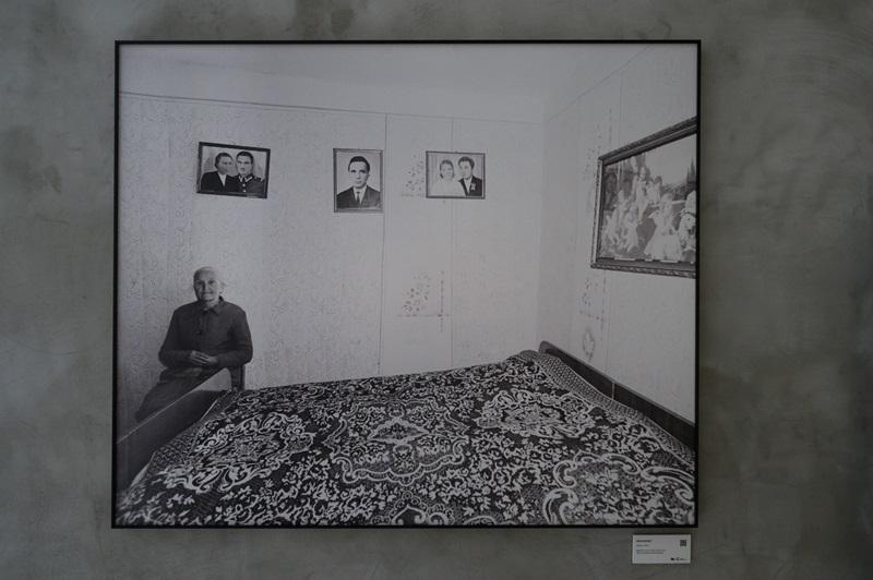 Fotografia autorstwa Zofii Rydet wyeksponowana na ścianie w czarnej ramie. Na zdjęciu widać starszą kobietę siedzącą w pokoju, obok dużego łóżka z wzorzystą narzutą. Na ścianach pokoju wiszą zdjęcia.