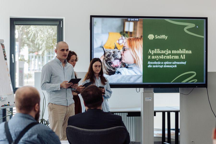 Troje członków zespołu Sniffly prezentuje swój projekt. Na slajdzie zdjęcie psa i napis: „Sniffly. Aplikacja mobilna z asystentem AI. Rewolucja w opiece zdrowotnej dla zwierząt domowych”