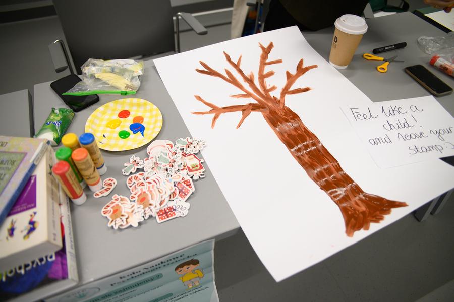 Materiały plastyczne na stoisku Koła Naukowego Psychologii Dziecka. Kartka z namalowanym drzewem, kolorowe farby i karteczki z obrazkami świątecznymi