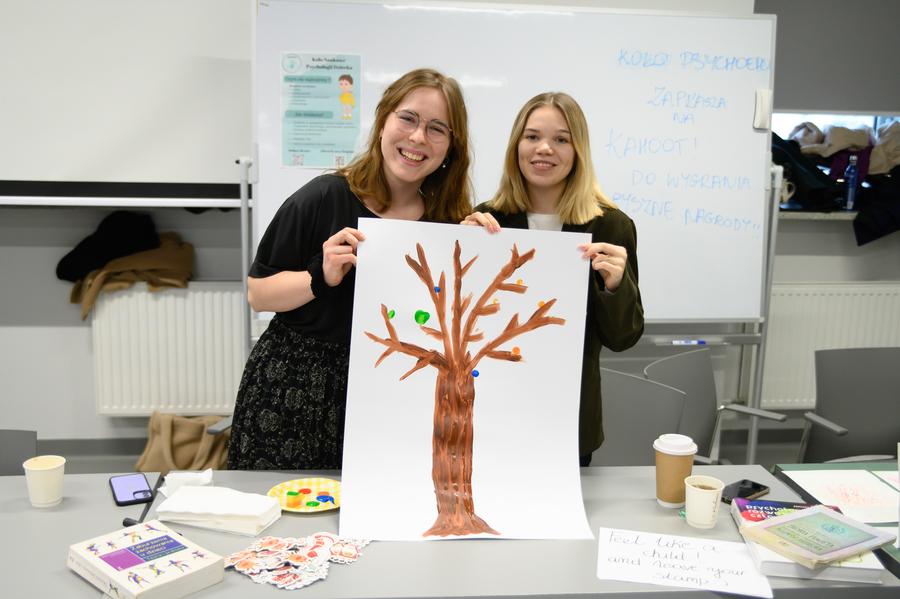 Członkinie Koła Naukowego Psychologii Dziecka trzymają kartkę z namalowanym drzewem, któremu uczestniczki i uczestnicy wydarzenia domalowali kolorowe listki