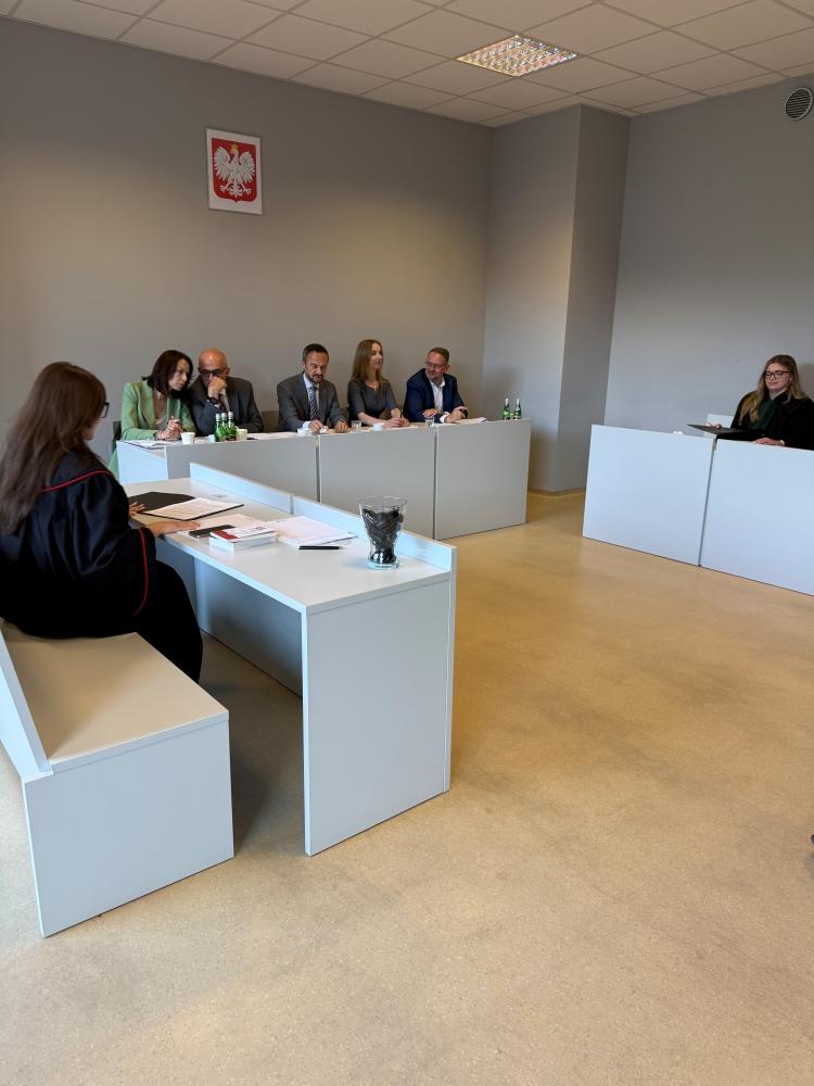 Po lewej stronie przy biurku siedzi uczestniczka konkursu, w tle widać jury.