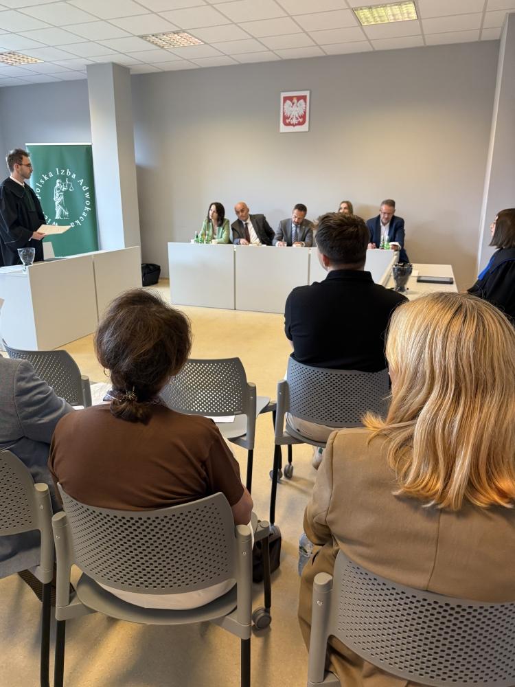 Na pierwszym planie publiczność na krzesełkach, po lewej stronie stoi uczestnik konkursu, w głębi sali jury siedzi za biurkiem.