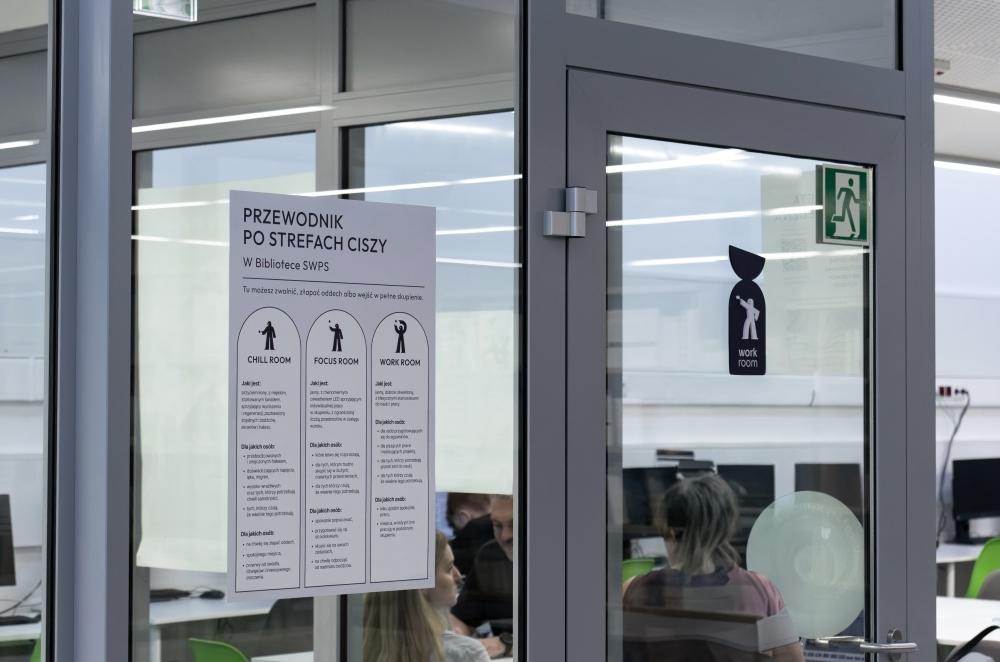 Plakat „Przewodnik po strefach ciszy” na szklanej ścianie oraz czarna tabliczka z piktogramem i napisem „work room” na drzwiach do sali w bibliotece.