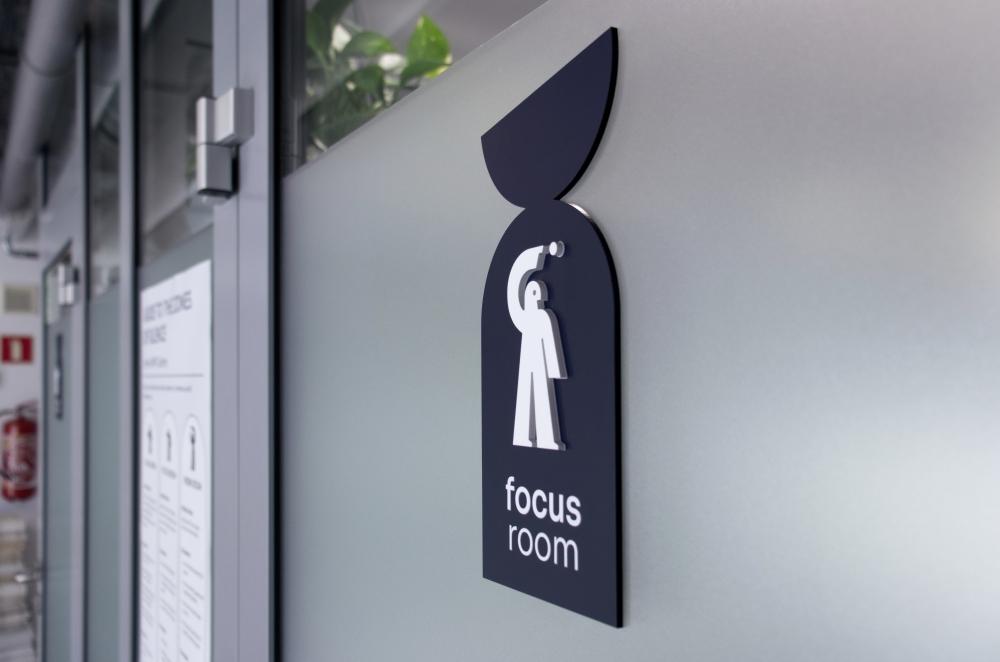 Widok z boku na ciemnoniebieską tabliczkę na szybie z białym piktogramem postaci trzymającej kropkę nad głową oraz napisem „focus room”.