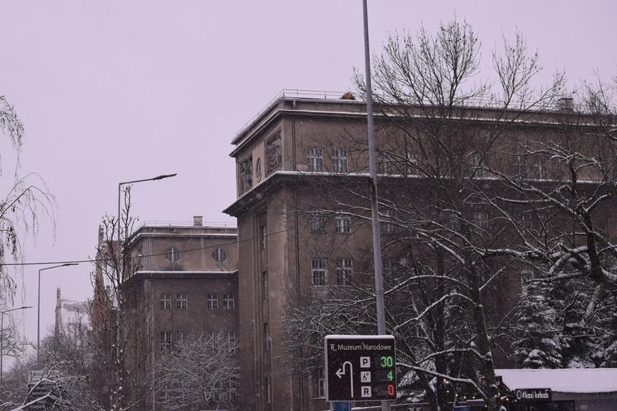 Widok Seminarium Śląskiego ze znajdującej się w pobliżu jedni. Widać dwa skrzydła budynku, ośnieżone drzewa, kawałek przystanku autobusowego i fragment znaku drogowego, który wskazuje kierunek do Muzeum Narodowego. 