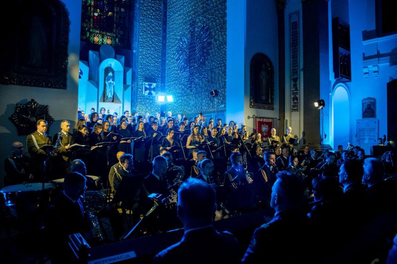 Chór Akademicki Uniwersytetu SWPS śpiewa „Requiem” w Katedrze Polowej Wojska Polskiego w Warszawie. Na pierwszym planie zdjęcia widać pierwszy rząd widowni, odwróconej plecami. Za nimi stoją orkiestra i chórzyści, oświetleni żółtym światłem