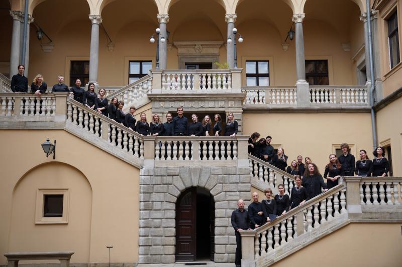 Zdjęcie grupowe około 35 osób. Chórzystki i chórzyści pozują do zdjęcia na schodach od strony dziedzińca w oficjalnych czarnych strojach, opierając się o barierki schodów