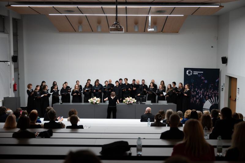 Zdjęcie grupowe chórzystów w Auli Tomaszewskiego w warszawskim kampusie Uniwersytetu SWPS. Chór śpiewa, odwrócona do nas plecami dyregentka dyryguje, publiczność siedzi w ławkach