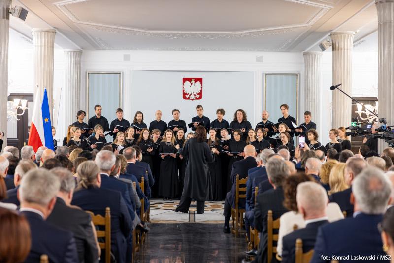 Sala Kolumnowa w Sejmie. Chór występuje przed licznie zgromadzoną widownią. Po lewej stronie flaga Polski i Unii Europejskiej, na ścianie na środku wisi godło Polski