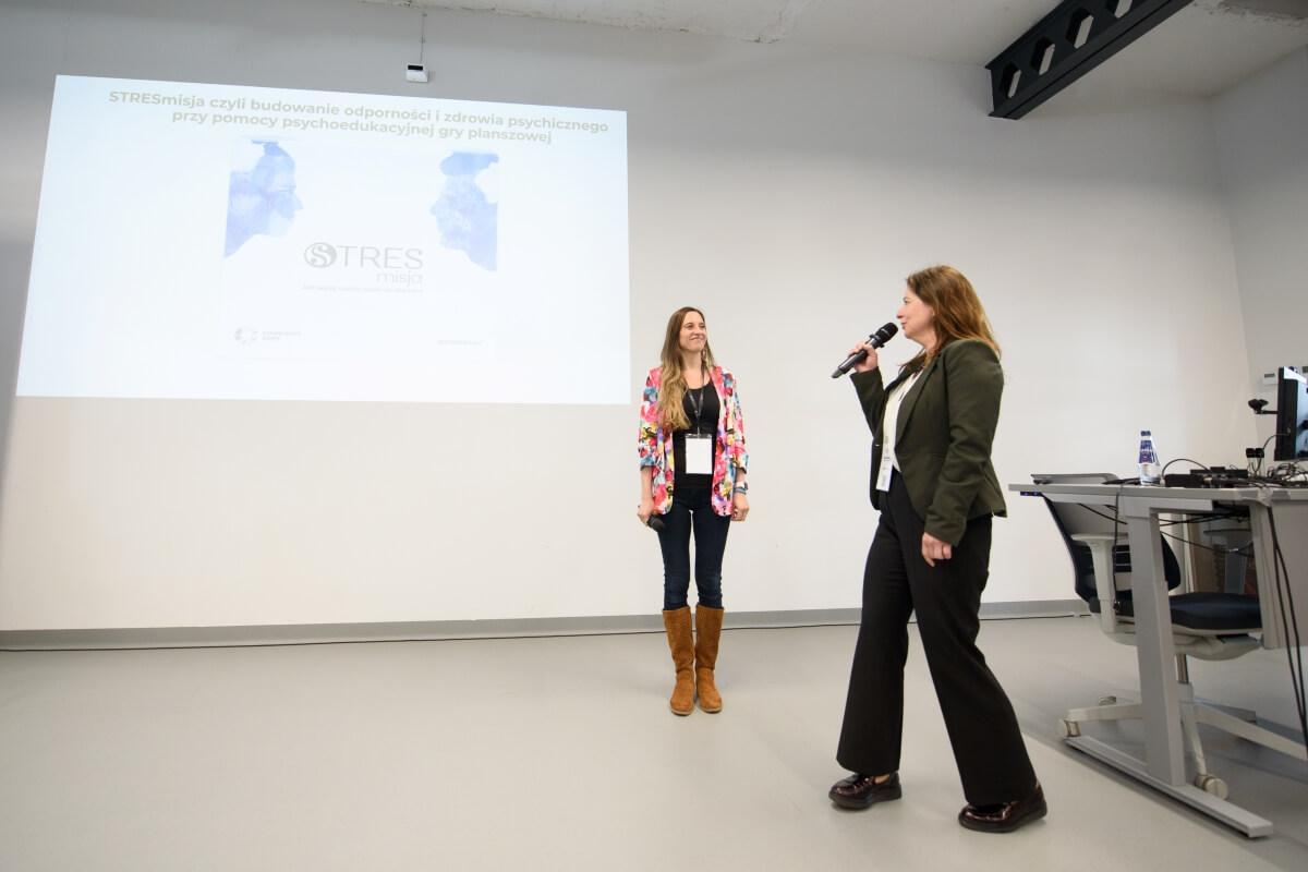 Dr Edyta Bonk i Katarzyna Barcińska z Uniwersytetu SWPS prezentują edukacyjną grę planszową STRESmisja podczas konferencji "Innowacje społeczne. Wyzwania-rozwiązania-praktyki"