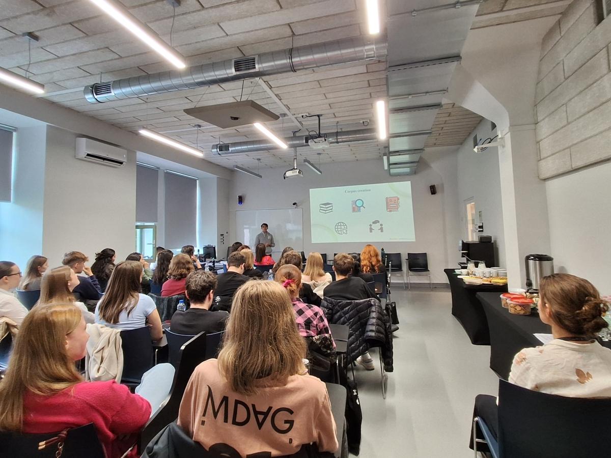 Uczestnicy konferencji widziani tyłem; siedzą w rzędach i obserwują prelegenta w głębi sali, który prezentuje slajdy.