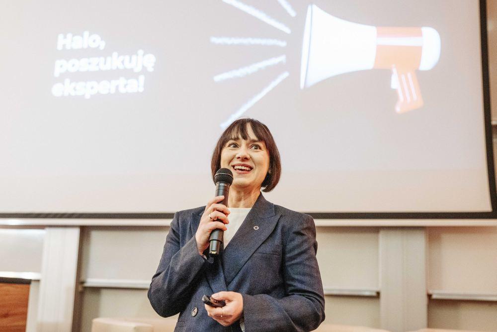 Renata Czeladko mówi do mikrofonu. W tle slajd z napisem „Halo, poszukuję eksperta!”