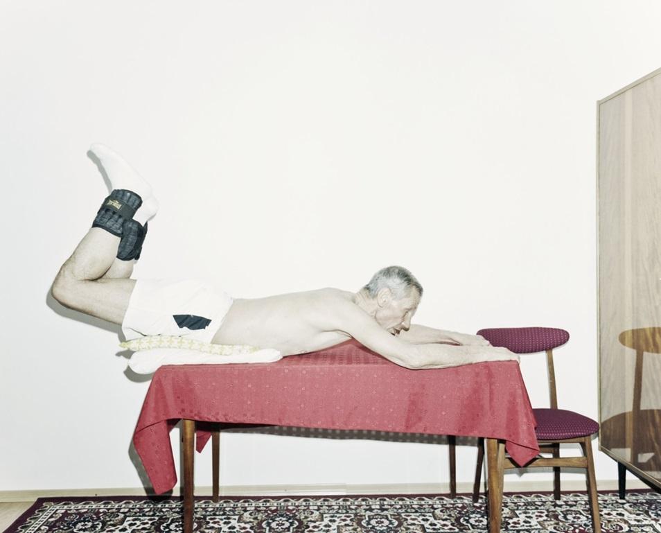 Zdjęcie przedstawiające starszego mężczyznę bez koszulki, w sportowych spoednkach, butach i skarpetkach, leżącego na stole przykrytym obrusem, w pokoju w stylu lat osiemdziesiątych. 